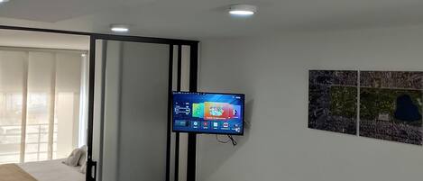 A hotel room with a bed, a bedside table, a TV mounted on the wall, and a window with curtains.