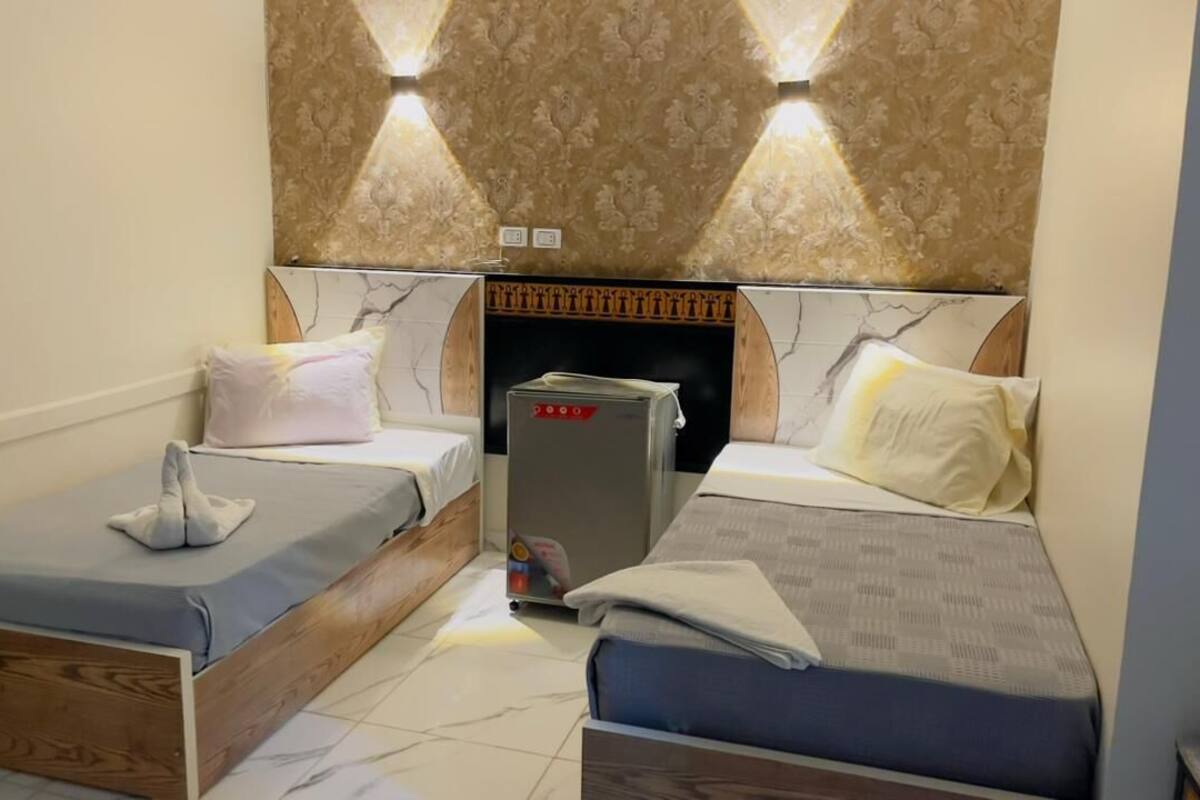 A hotel room with two beds, a small table, a minibar, and a decorative wall.