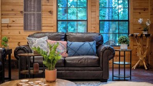 A cozy cabin interior with a leather sofa, a small coffee table, and a window with a view of trees. - Close to All~Mini Golf~Fire-Pit~Swing Set~Hot Tub (Broken Bow)