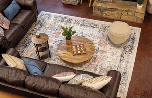 A cozy living room with a brown leather sofa, a round wooden coffee table with a plant and wooden blocks spelling "XOXO," and a patterned rug.