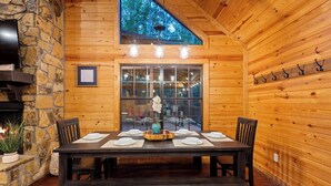 A wooden interior with a stone fireplace, a dining table set for four, and a large window overlooking a forest. - Close to All~Mini Golf~Fire-Pit~Swing Set~Hot Tub (Broken Bow)