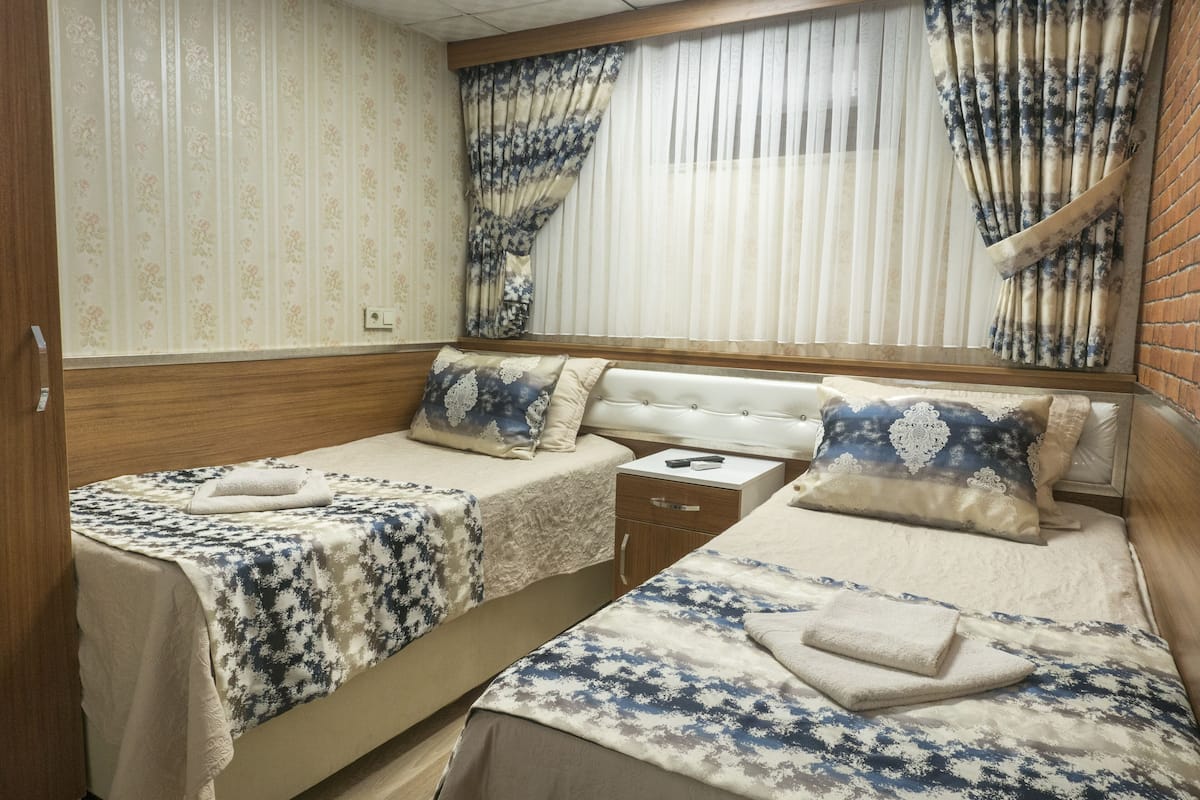A double room with two beds, each with a patterned bedspread and pillows, a wooden headboard, a small bedside table, and a window with curtains.
