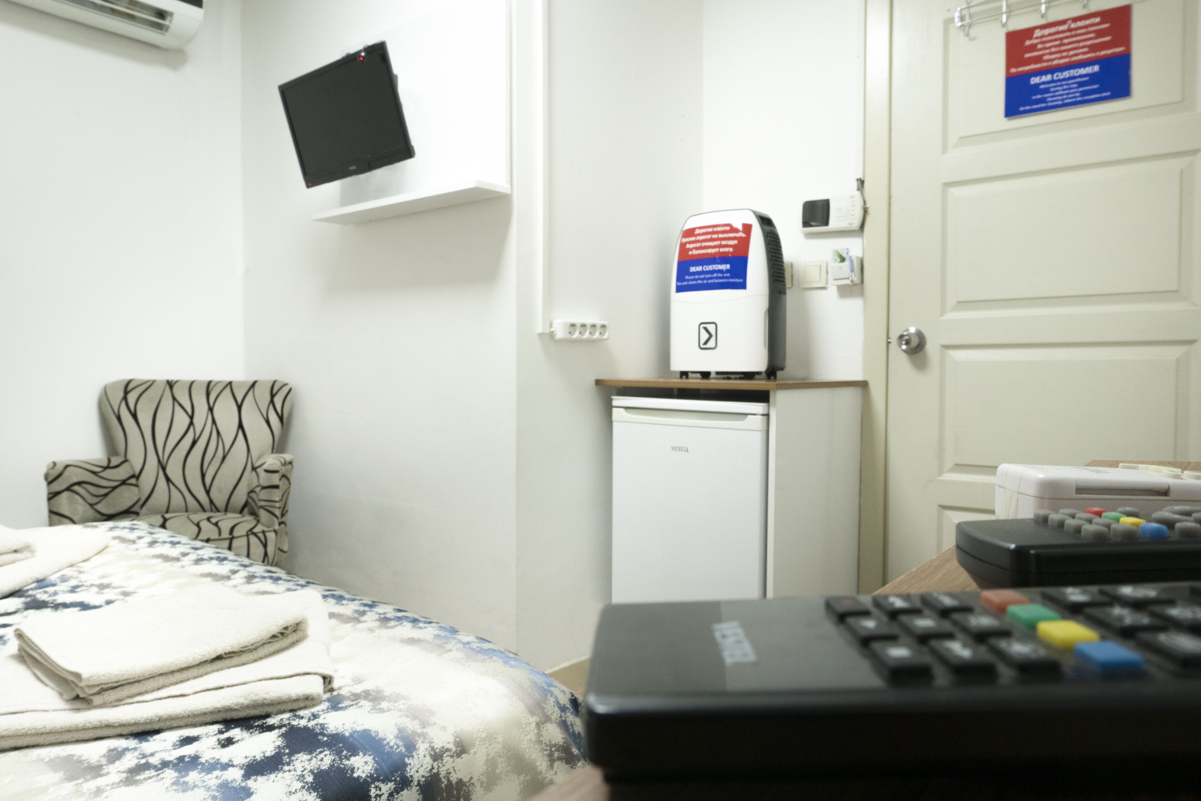A hotel room with a bed, a chair, a TV mounted on the wall, a mini-fridge, and a control panel.