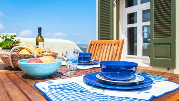 A table set with blue plates, bowls, and a wine bottle, with a view of the sea and a building with green shutters in the background. - Beautiful calm rooftop studio with stunning views (CHANIA)