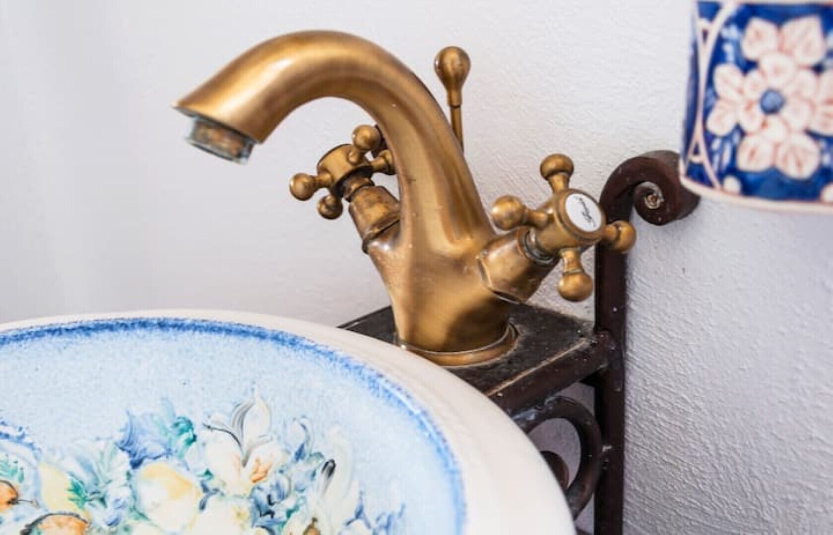A vintage brass faucet mounted on a wall, a decorative ceramic sink with floral patterns, and a blue and white ceramic container with a floral design.
