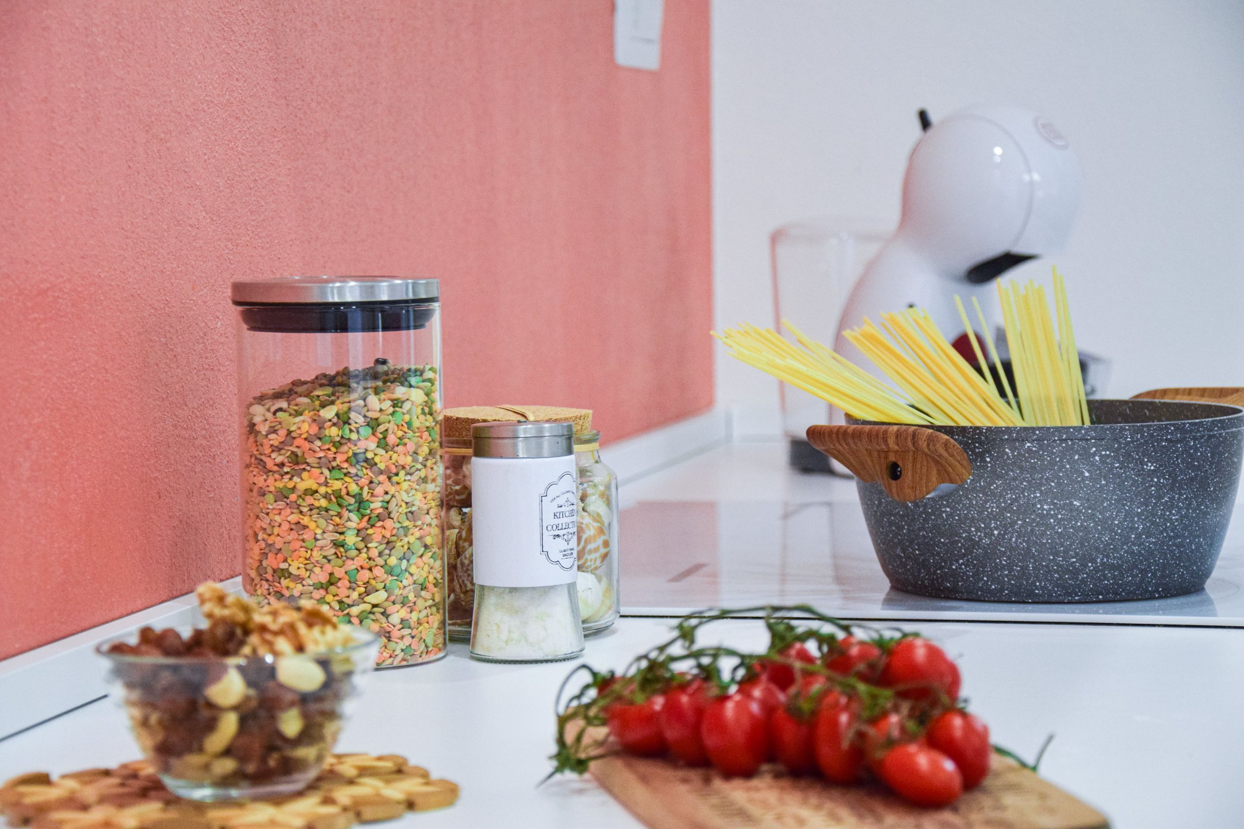 Un plan de travail de cuisine avec des bocaux d’épices, un bol de noix, une casserole de spaghettis et une barquette de tomates cerises.