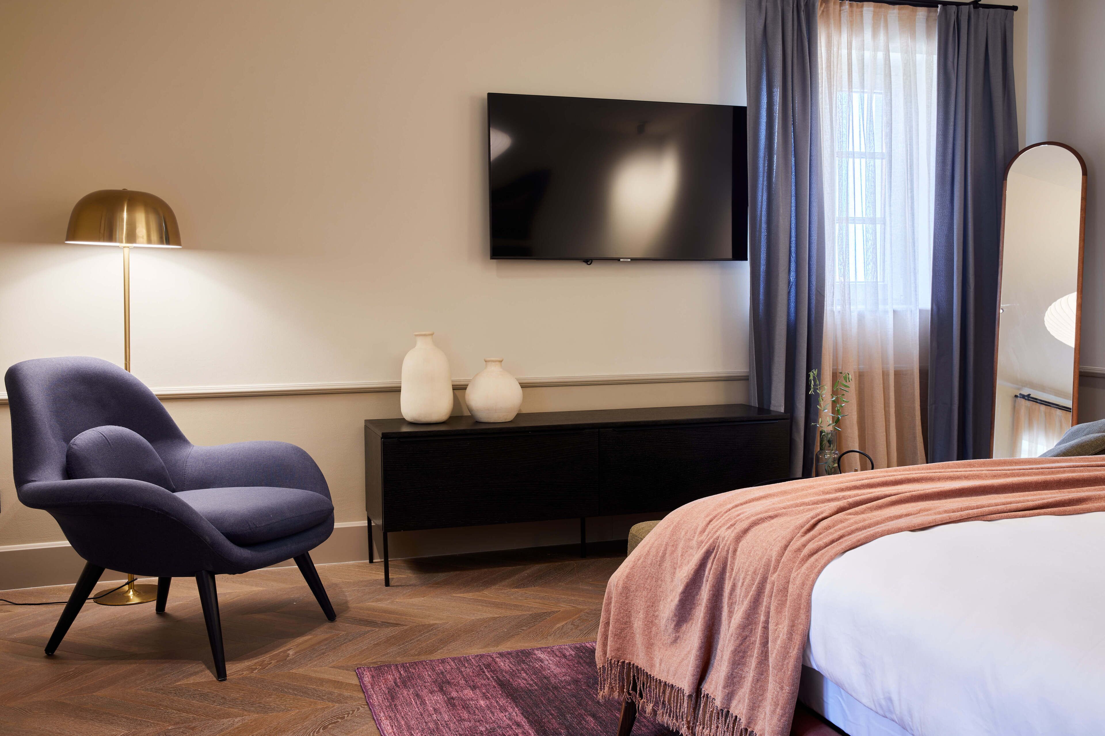 A modern hotel room with a bed, a flat-screen TV mounted on the wall, a dark-colored console table with decorative vases, a blue armchair, and a large mirror.