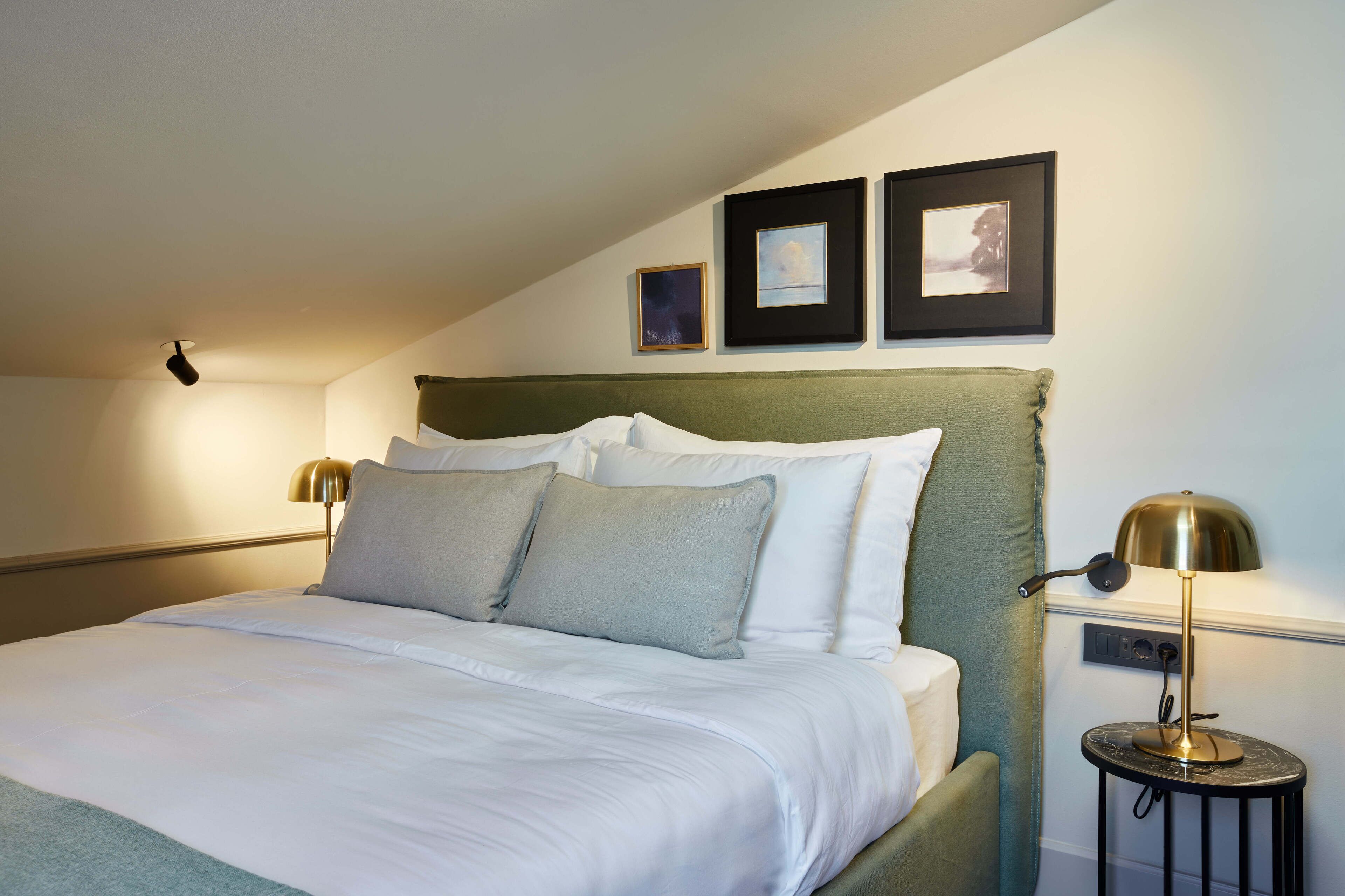 A neatly made bed with a green headboard, two bedside lamps, and two framed pictures on the wall.