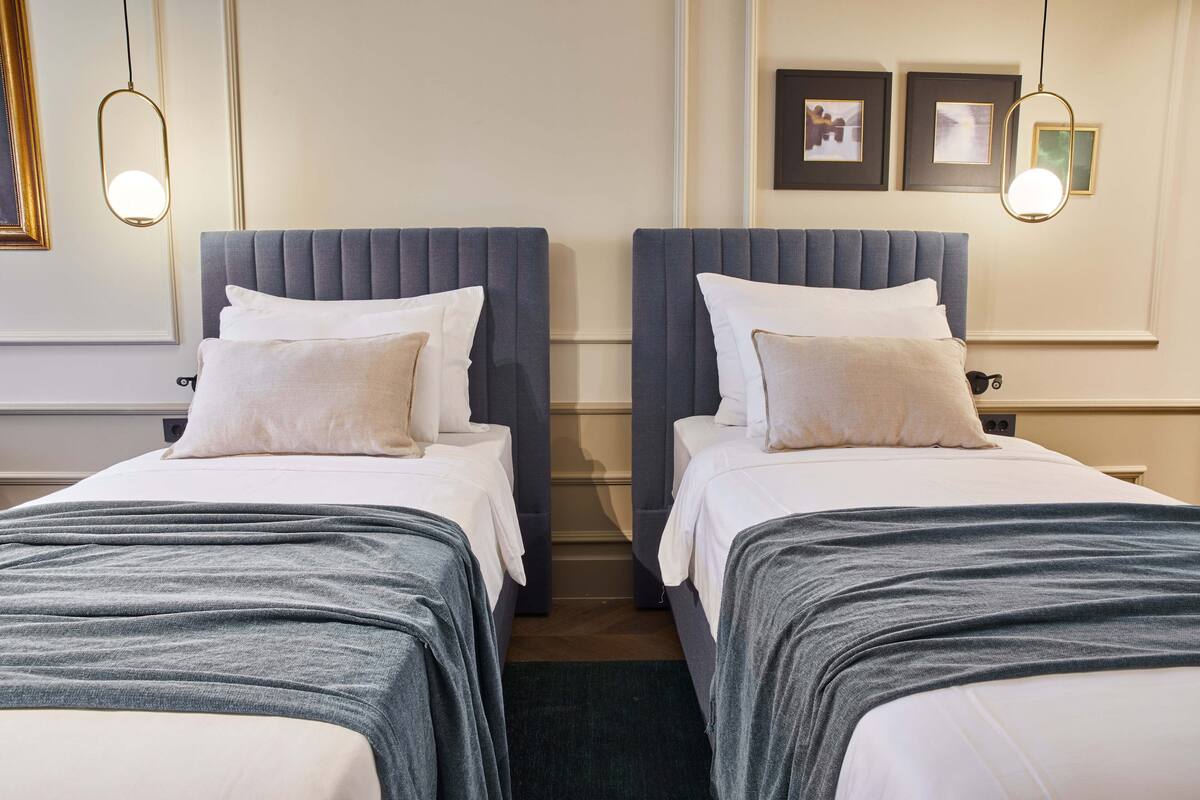 Une chambre d’hôtel avec deux lits, chacun doté d’une tête de lit grise, de draps blancs et d’un plaid gris. Deux lampes suspendues sont accrochées au-dessus des lits.