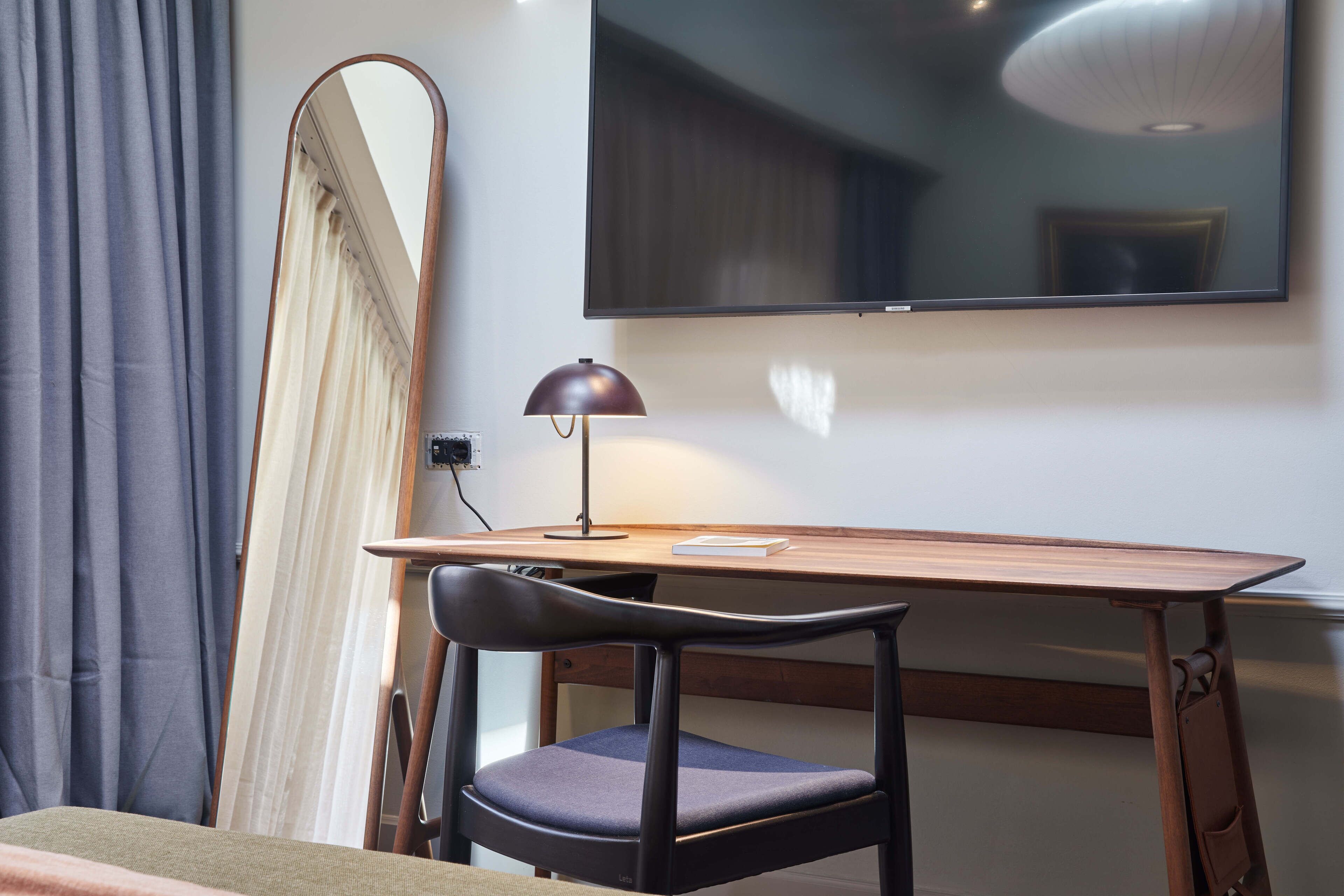 Ein Zimmer mit Holzschreibtisch, Stuhl, Lampe, Spiegel und einem an der Wand befestigten Fernseher.