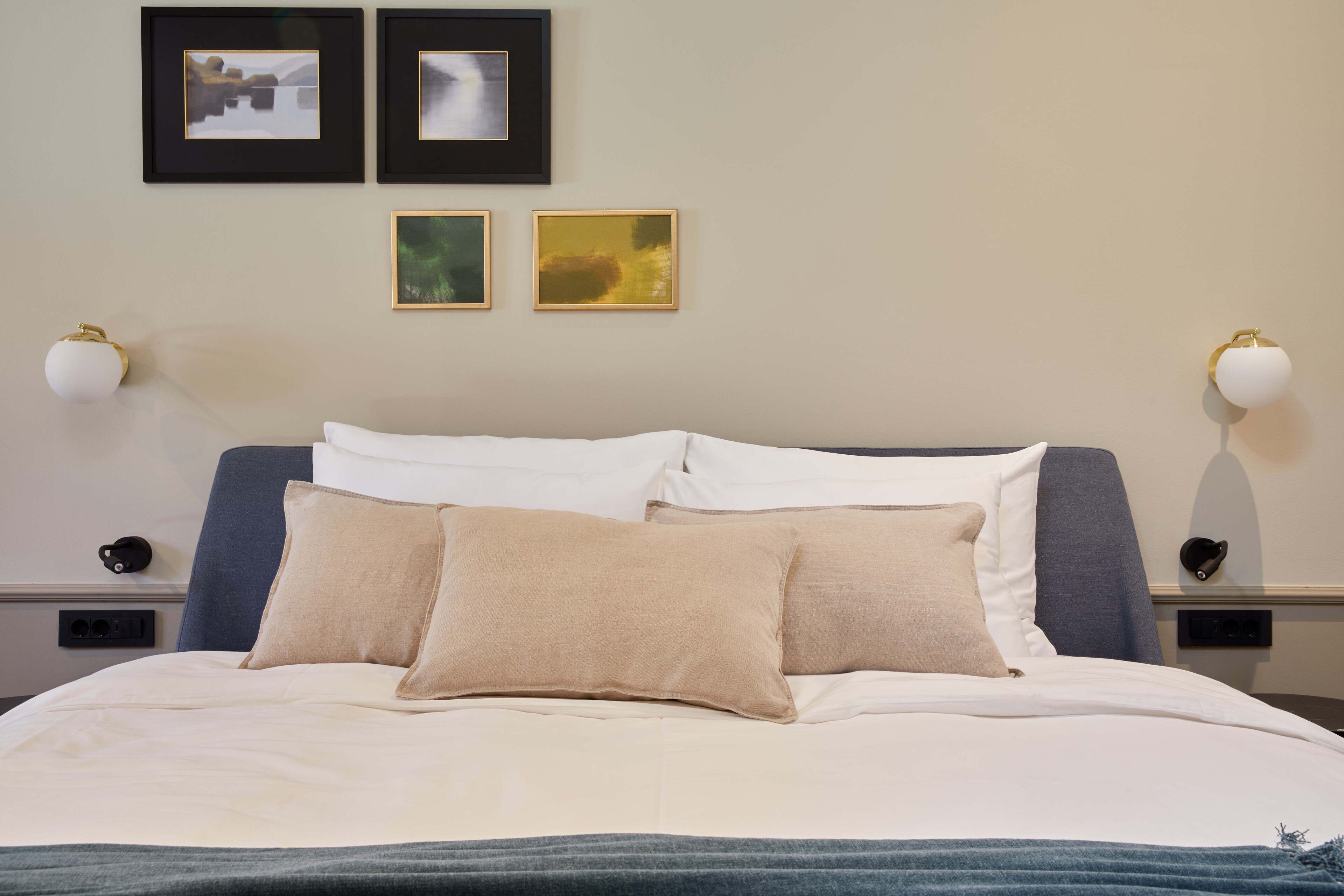 Cama bem arrumada com roupa de cama branca e bege, duas molduras na parede e duas luminárias de parede.