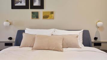 Cama bem arrumada com roupa de cama branca e bege, duas molduras na parede e duas luminárias de parede.