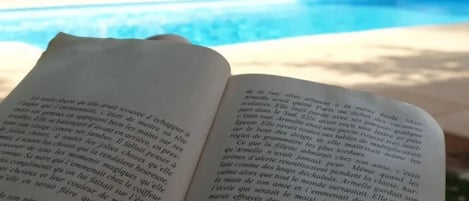 An open book held by a hand with a pool and trees in the background.