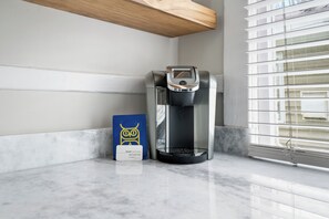 A coffee machine on a marble countertop next to a blue bag with a logo, in a room with a window and blinds.