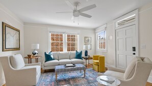 A living room with a white sofa, two armchairs, a coffee table, and a patterned rug. There are two windows with white frames, a ceiling fan, and a door with a white frame.