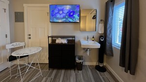 A compact hotel room with a flat-screen TV, a small white table, a black cabinet, a sink, and a window with curtains.