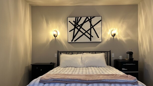 A neatly made bed with striped bedding, a black abstract painting above it, and two wall-mounted sconces on either side.