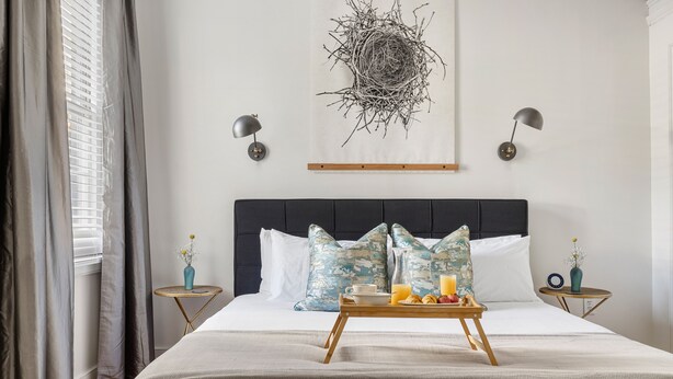 A neatly made bed with a tray of breakfast items, a wall-mounted abstract art piece, and two wall sconces. - The Feather | Next To Bad Bunnies Coffee (Charleston)