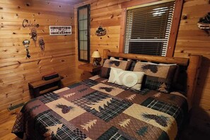 Ein gemütliches Schlafzimmer im Blockhausstil mit Holzvertäfelung, einem Bett mit Decke, einem Nachttisch, einer Lampe und einem Fenster mit Jalousien.
