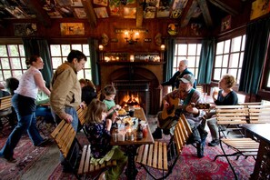 Un pub acogedor con muebles de madera, una chimenea y un ambiente animado con gente cenando y un músico tocando la guitarra.