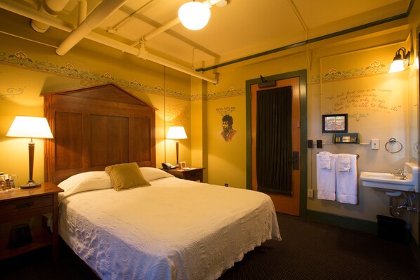 Una habitación de hotel con una cama grande, dos lámparas de noche, un cabecero de madera, una pared con un borde decorativo, una puerta y un baño con lavamanos y toallas.