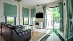 Un salon moderne avec un canapé en cuir, une table basse en verre et un téléviseur à écran plat fixé au mur. Une porte-fenêtre coulissante donne accès à un espace extérieur avec vue sur la verdure.