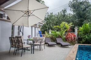 Un patio al aire libre con una mesa y sillas, una silla de descanso, una piscina y abundante vegetación.