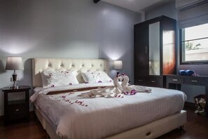 Una cama bien hecha con sábanas blancas y pétalos de rosa, una lámpara de noche y un armario de madera oscura.