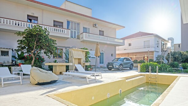 Una piscina con sillas de descanso y una escalera, rodeada por un edificio con balcones y un coche estacionado.