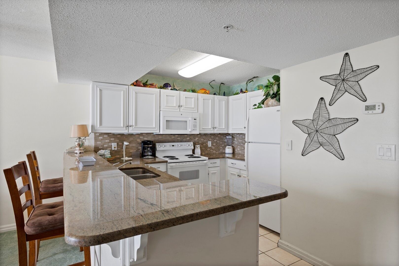 Une cuisine avec des armoires blanches, un comptoir en granit et un pare-feu orné d’une fresque tropicale. Deux décorations en forme d’étoile de mer sont accrochées au mur.