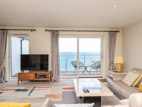 A modern living room with a television, sofa, coffee table, and a balcony with a view of the sea.