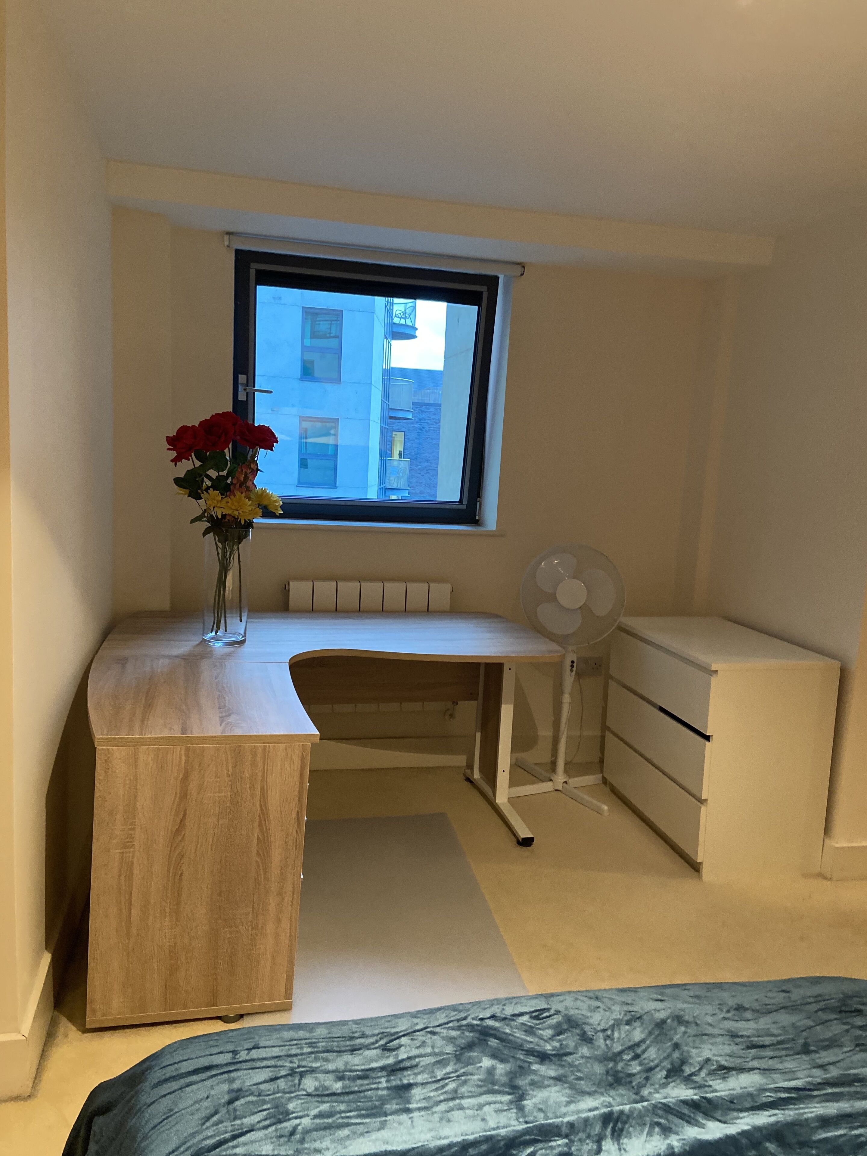 A bedroom with a wooden desk, a radiator, a window, a fan, and a bed.