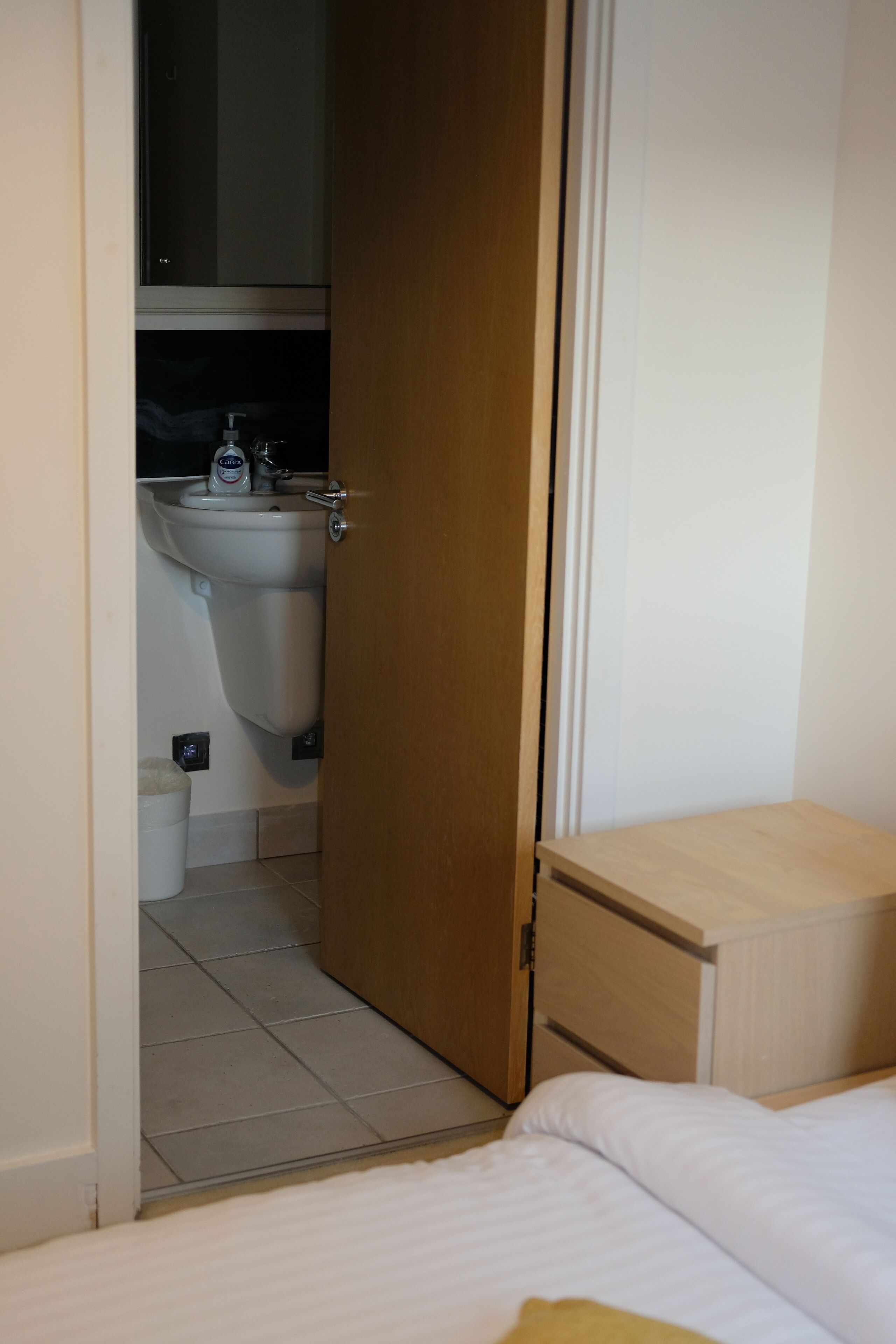 Une chambre à coucher avec un lit, une table de chevet en bois et une salle de bain visible à travers une porte ouverte.