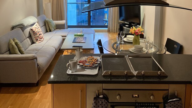 Een moderne keuken met een centraal kookeiland, een eethoek met een glazen tafel en een zithoek met een bank.