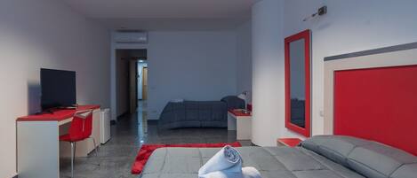 A modern hotel room with a bed, a desk with a computer, a TV, and a red-framed mirror.