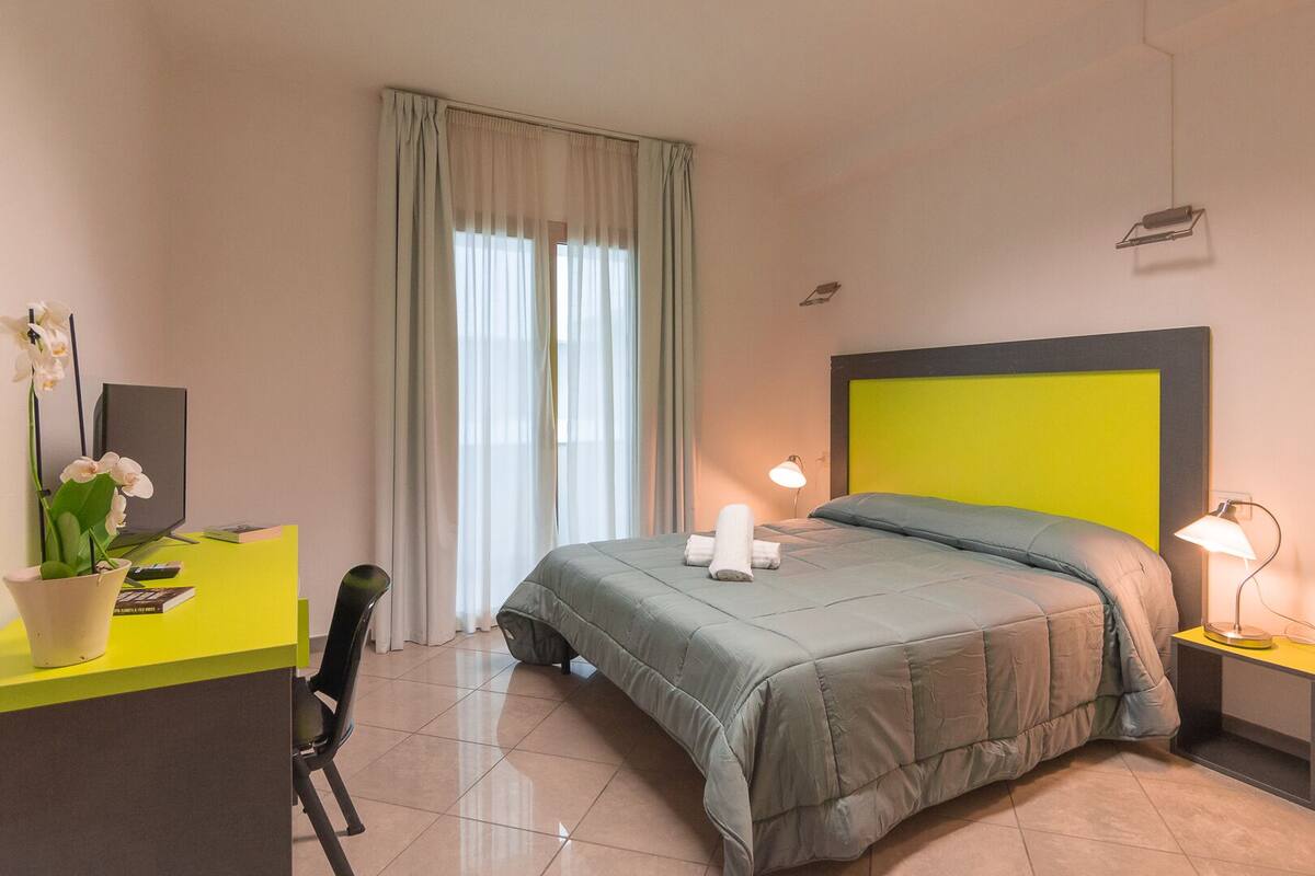 A hotel room with a bed, a desk, a chair, a TV, and a window with curtains.