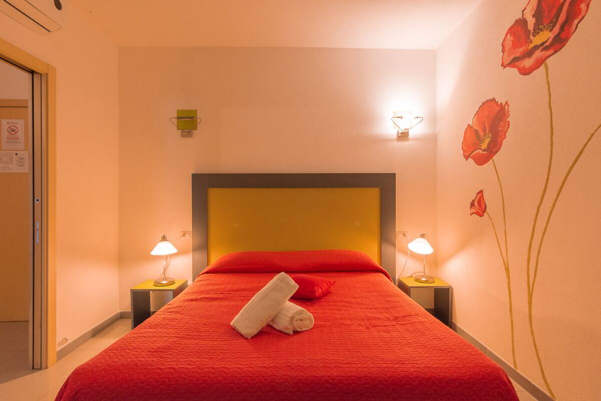 A hotel room with a red bedspread, two bedside lamps, a towel on the bed, and a wall with a floral design.
