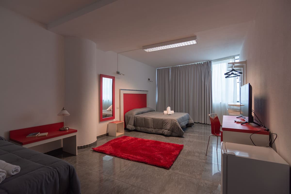 A modern hotel room with a bed, a desk with a computer, a red rug, and a TV.
