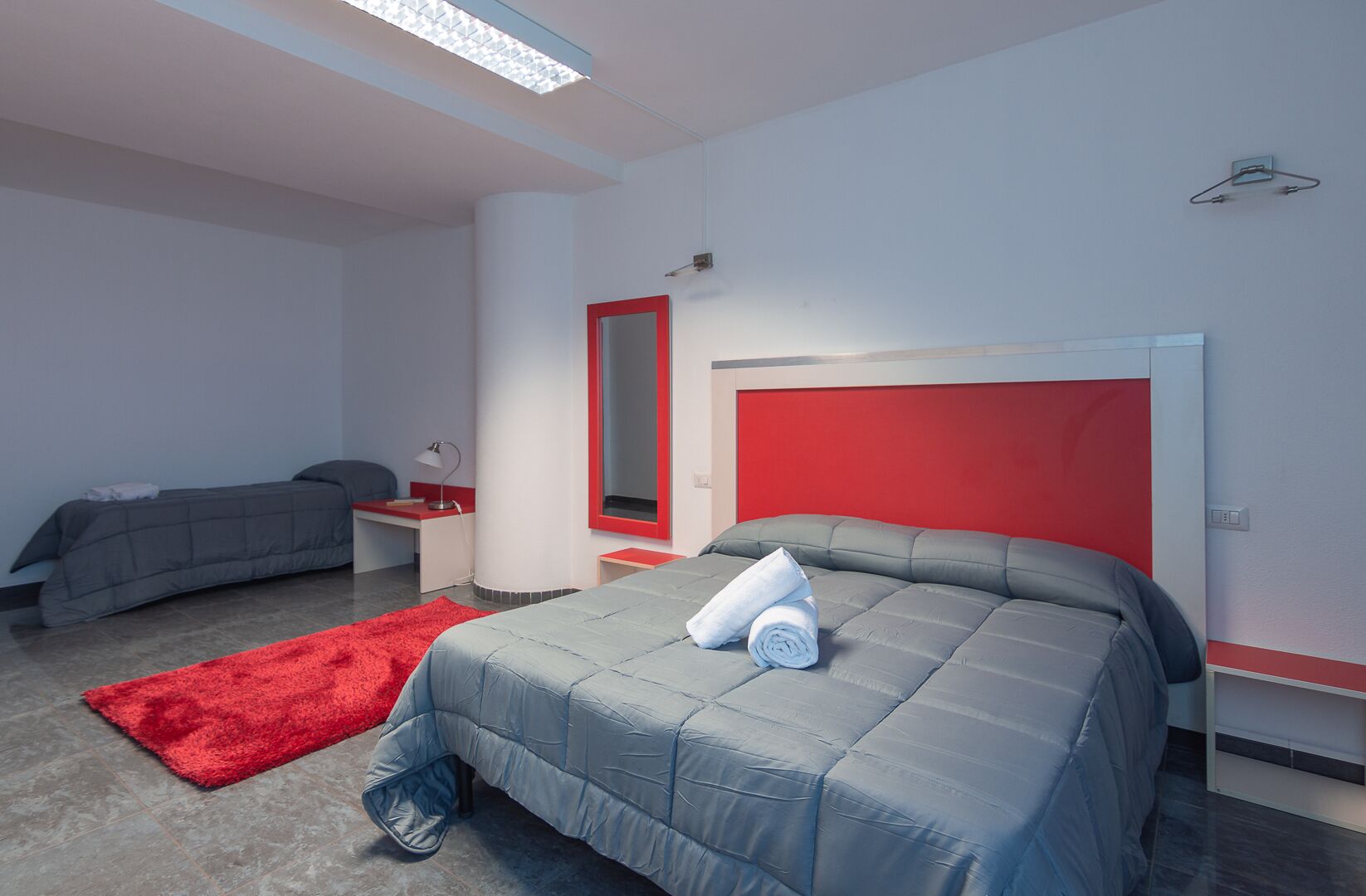 A modern hotel room with a large bed, a smaller bed, a red accent wall, and a red rug.