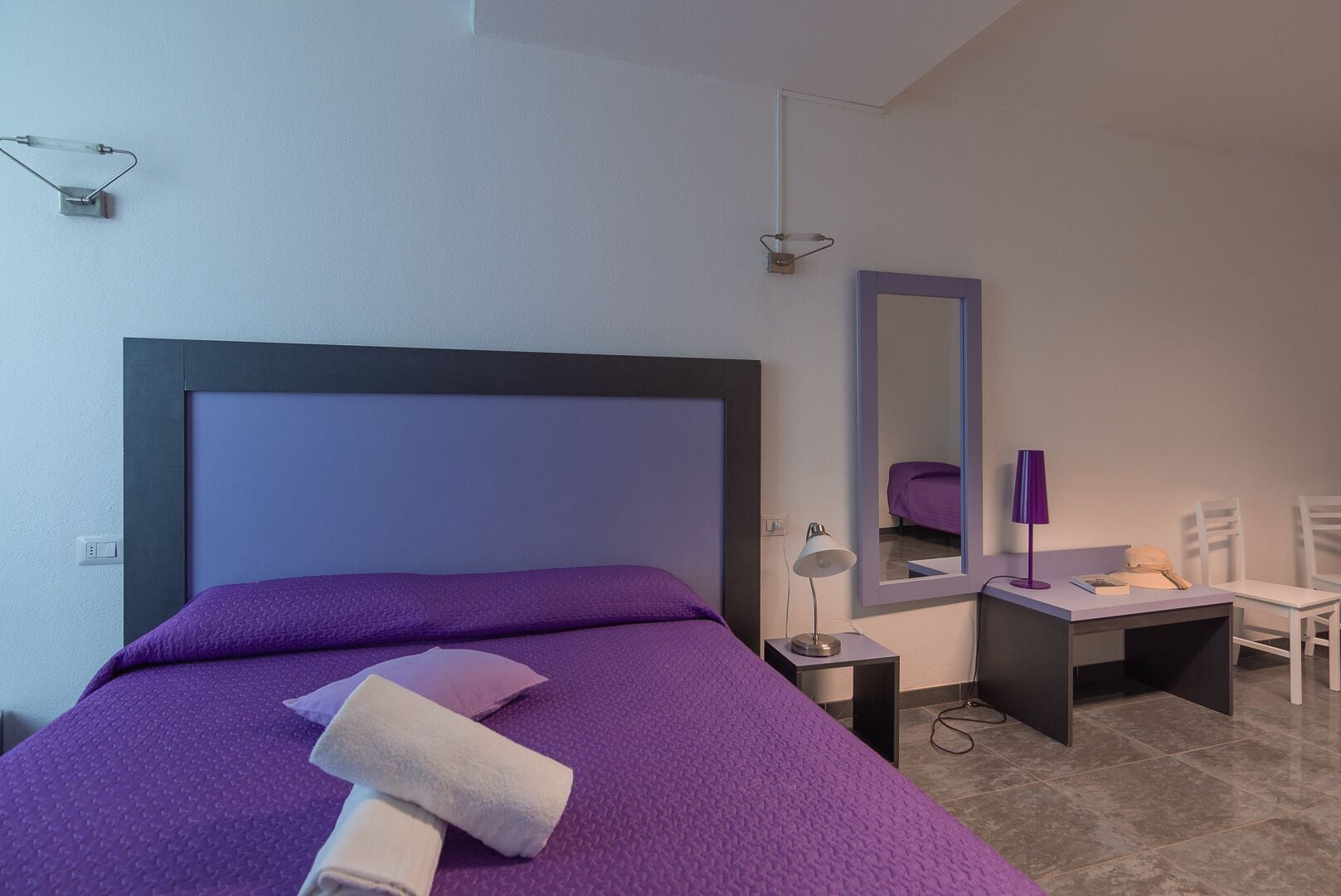 Un dormitorio moderno con una cama grande, una mesita de noche, un escritorio, una silla, un espejo y una lámpara.