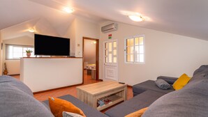 A modern living room with a flat-screen TV, grey sofas, a wooden coffee table, and a small kitchen area in the background. - Sunset House (Arco da Calheta)
