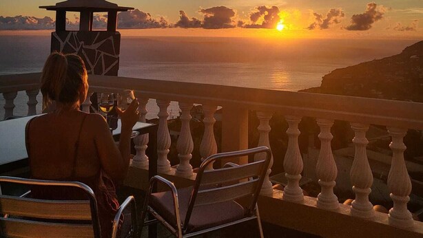 A balcony with a view of the sunset over the sea, a small gazebo, and a person seated at a table. - Sunset House (Arco da Calheta)