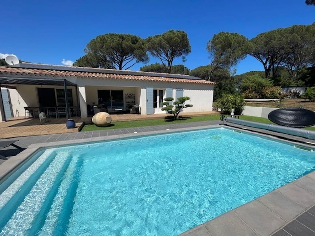 A swimming pool with a tiled deck, a covered patio area, and a house with a tiled roof and blue shutters.