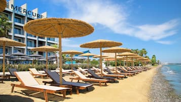 A beachside hotel with rows of sun loungers and thatched umbrellas, a clear blue sky, and a calm sea.