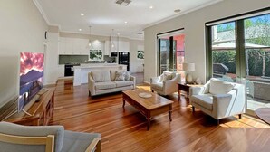 A spacious living area with a wooden floor, a sofa, a coffee table, and a TV. There is a kitchen in the background with white cabinets and a dining area with a table and chairs.