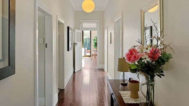 A hallway with wooden flooring, white walls, and a large mirror on the right. There is a vase with flowers on a table in the foreground.