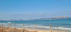 Una playa con una cerca de madera en primer plano, un cielo azul despejado y montañas a lo lejos.