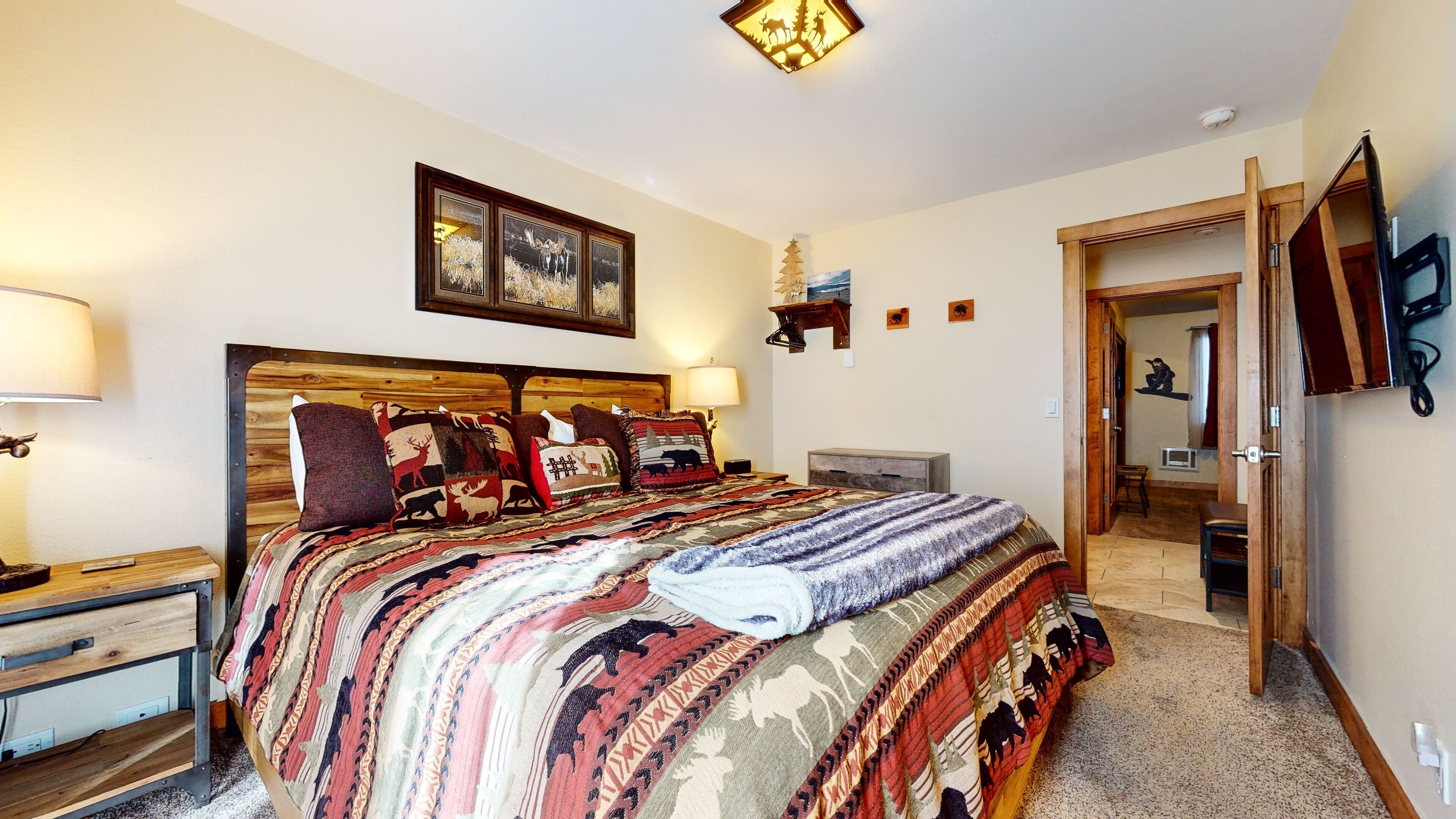 A bedroom with a large bed, wooden headboard, and multiple pillows with animal prints. There is a nightstand with a lamp, a small table, and a wall-mounted shelf with decorative items.