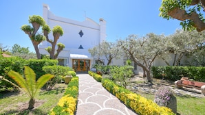 A pathway leading to a white building with a door, surrounded by well-maintained gardens and trees.