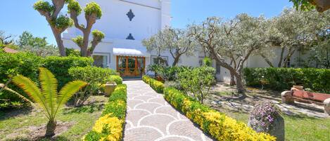 A pathway leading to a white building with a door, surrounded by well-maintained gardens and trees.